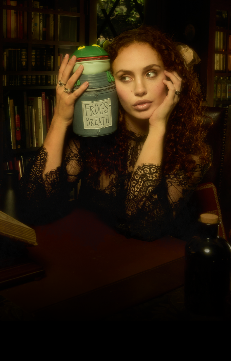 Model holding Frog's Breath Loungefly Crossbody bag up to her face and sitting in a creepy library setting.