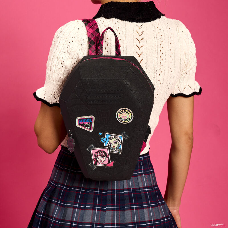 Woman wearing a white knitted sweater and plaid pleated skirt facing away from camera and wearing the coffin-shaped Monster High locker mini backpack. She stands against a pink background.