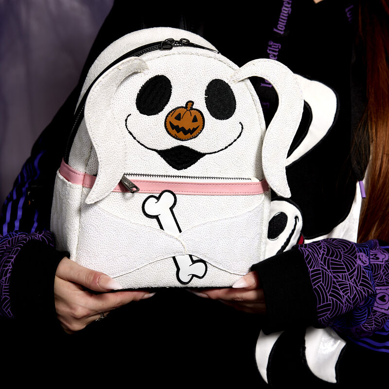 Woman holding the white sequin glow-in-the-dark Loungefly bag that looks like Zero from The Nightmare Before Christmas against a dark purple background.