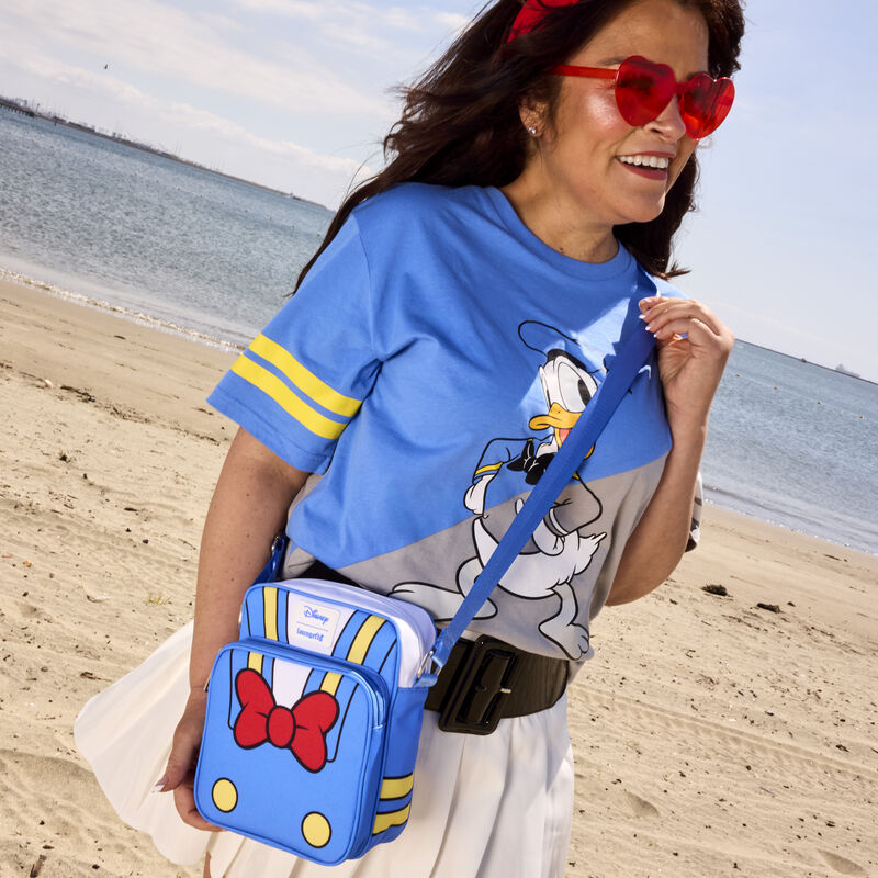 Woman standing on the beach in red sunglasses wearing the Loungefly Donald Duck 90th Anniversary Passport Crossbody Bag, featuring details of Donald's sailing uniform. 