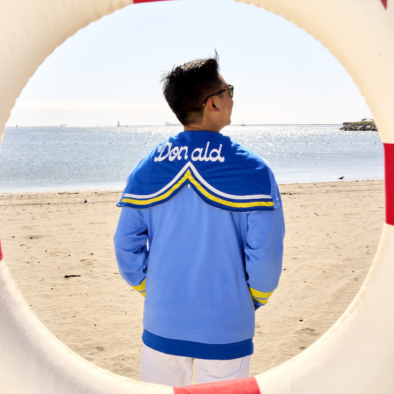 Man on the beach facing away from camera wearing the Loungefly Donald Duck 90th Anniversary Hoodie with the hood unzipped to show how it lays on the back of the hoodie as a collar that says "Donald"