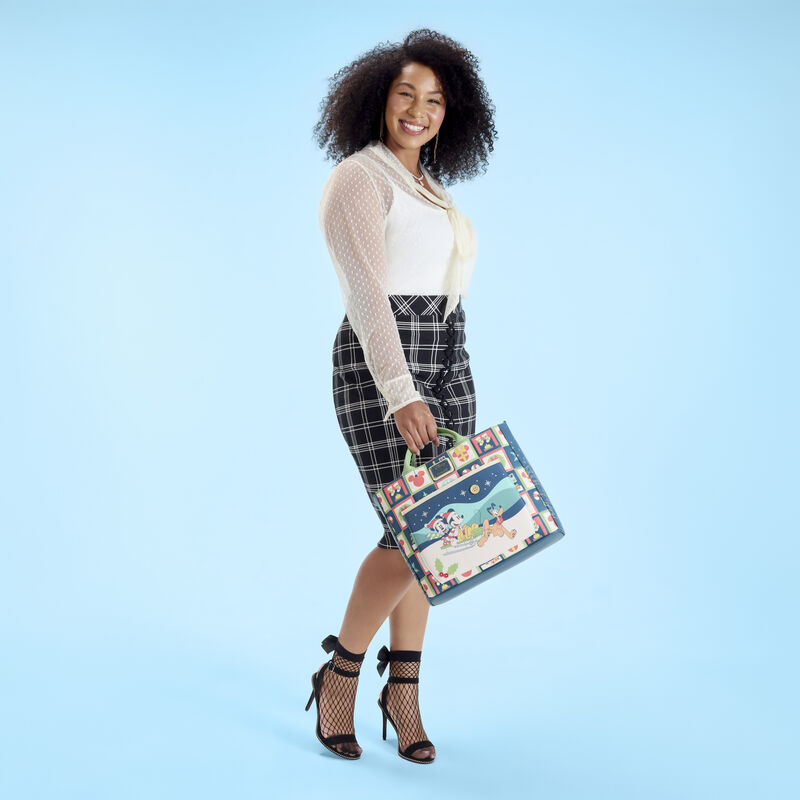 Woman wearing a black and white plaid skirt with a white blouse and black heels. She holds the Mickey & Friends Holiday Convertible Backpack & Tote Bag. She stands against a blue background. 