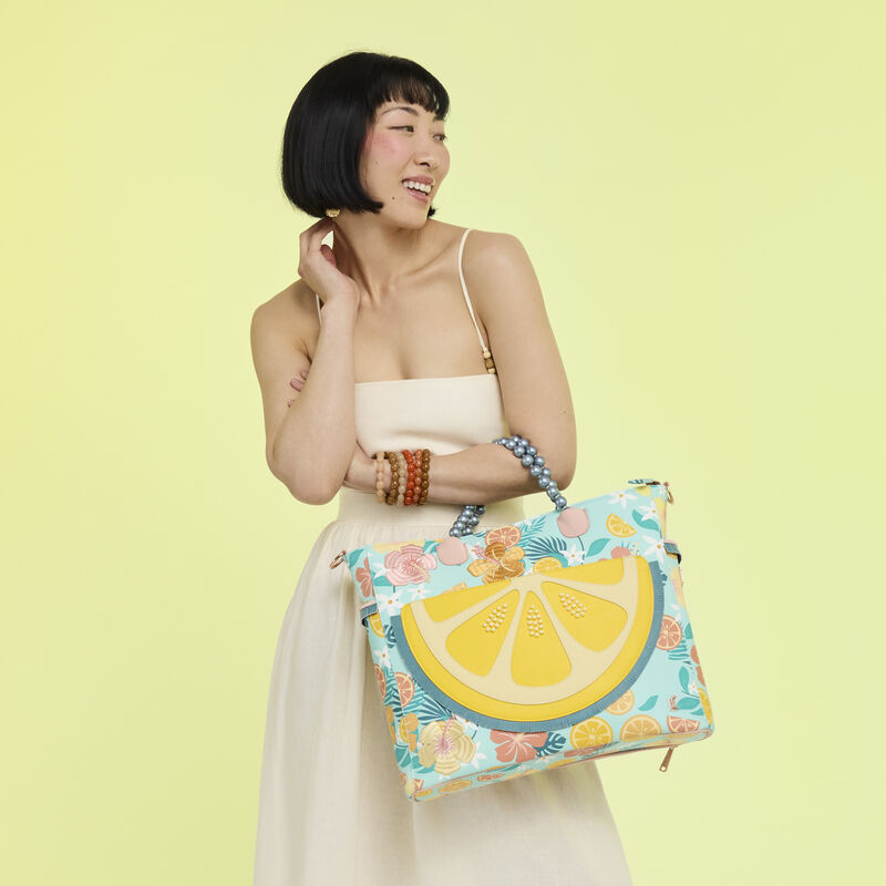 Woman wearing a white dress with the Loungefly Tropical Lemon Floral Convertible Backpack and Tote bag over one arm. She stands in front of a yellow background. 