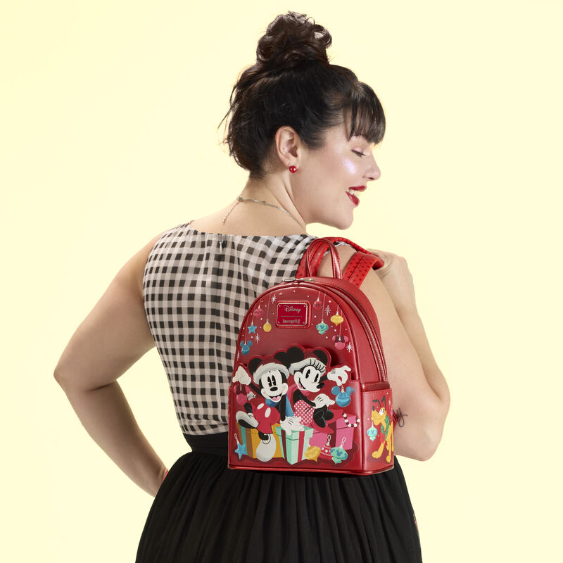Woman facing away from camera, wearing a black skirt and black-and-white checkered blouse. She wears the Loungefly Mickey and Friends Holiday Ornaments Mini Backpack over a shoulder. She stands against a yellow background. 