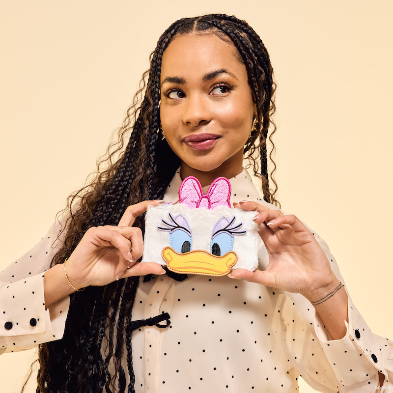 Woman facing camera wearing a sheer neutral black-polka-dot blouse and holding the sequin and plush Loungefly Daisy Duck Card Holder. She stands against a beige background. 