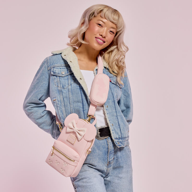 Woman smiling at camera wearing a white top, jeans, and a denim jacket. She wears the pink Loungefly Minnie Mouse Sling Crossbody. She stands against a pink background.