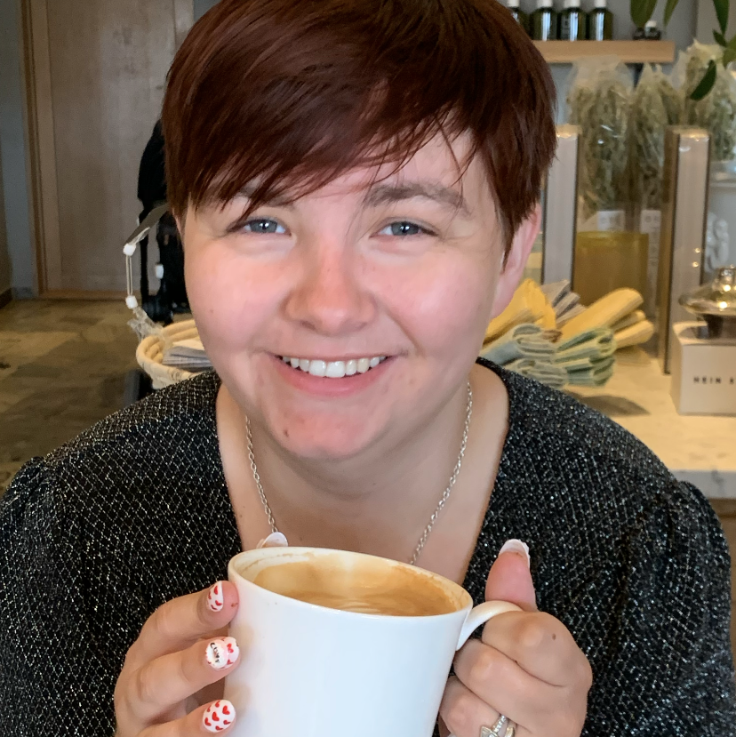 Image of our designer, Christina P., with short brown hair smiling at camera holding a cup of coffee. 