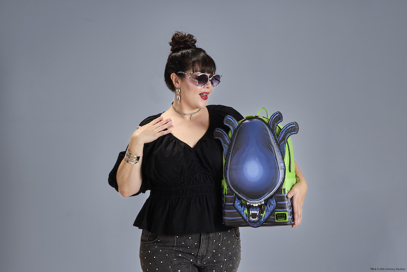 Woman wearing all black holding the Loungefly Alien Xenomorph full-size backpack. She stands against a grey background.