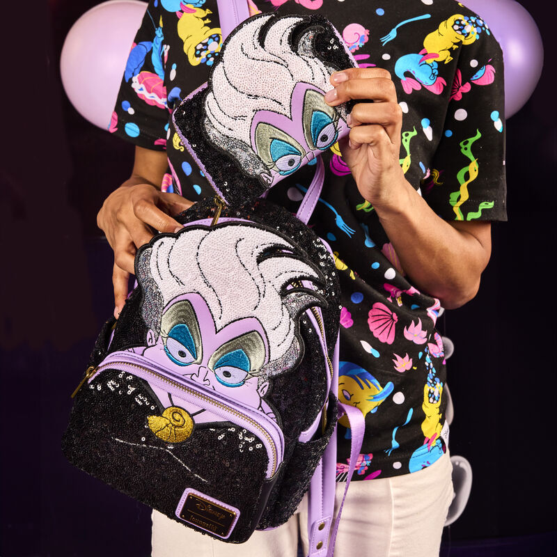 Woman facing the camera wearing a black shirt with neon Little Mermaid characters on it, holding the sequin Ursula mini backpack and the matching wallet. She's putting the Ursula Sequin wallet into the mini backpack. 