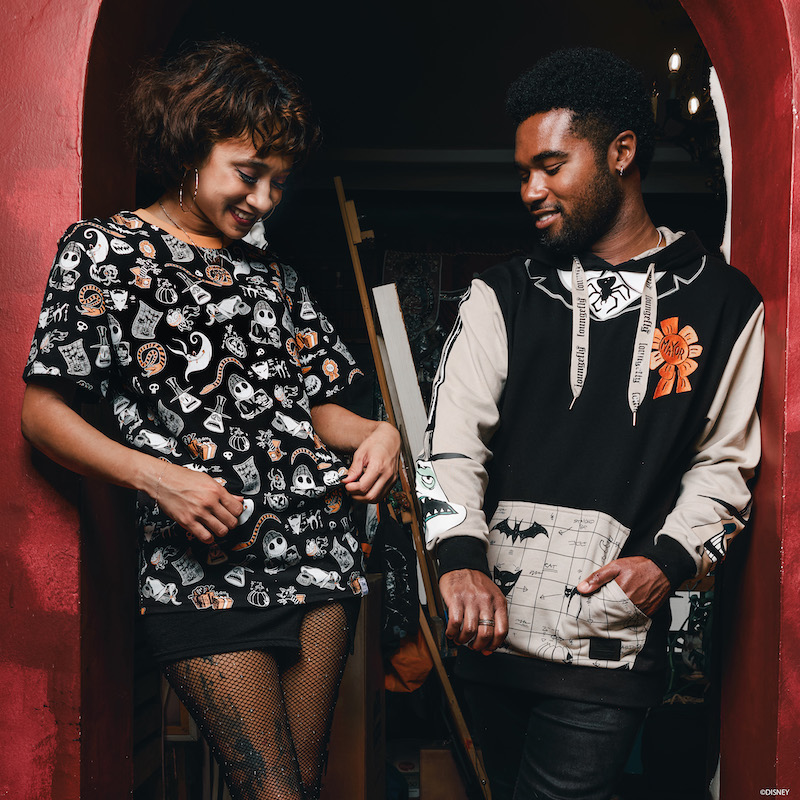 Woman and man standing together with stairs in the background. The woman wears a Nightmare Before Christmas all-over print tee shirt and the man wears a Loungefly hoodie inspired by the Mayor's outfit from Nightmare Before Christmas 