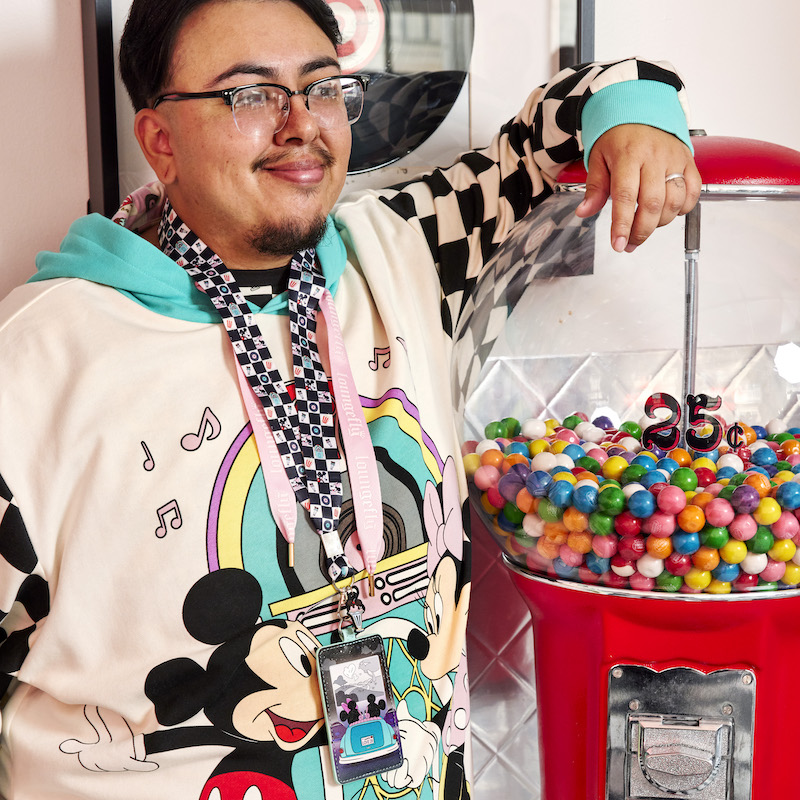 Image of man in the Date Night hoodie wearing the Mickey and Minnie Date Night Drive-In Lanyard with Card Holder standing beside a gumball machine.