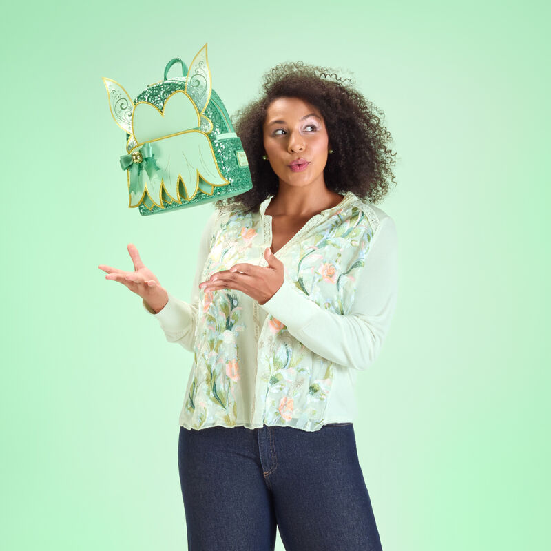 Woman wearing a green floral blouse and jeans, standing against a green background. She tosses the Loungefly Tinker Bell bag in the air to make it look like it’s flying. 