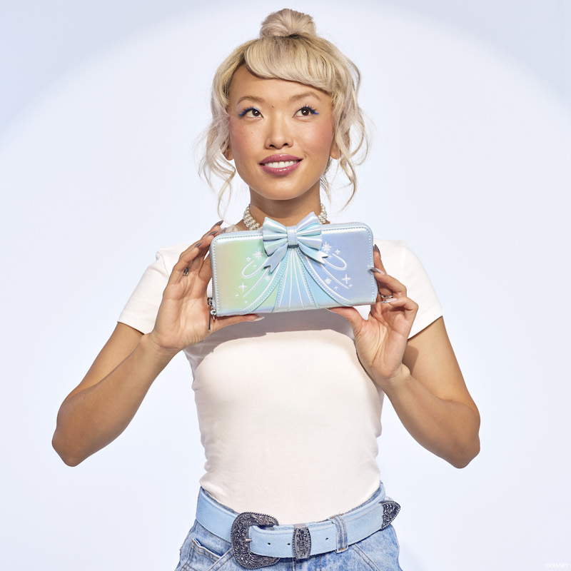 Blonde woman looking up and smiling and holding the Cinderella 75th anniversary Shiny Series iridescent wristlet wallet. 