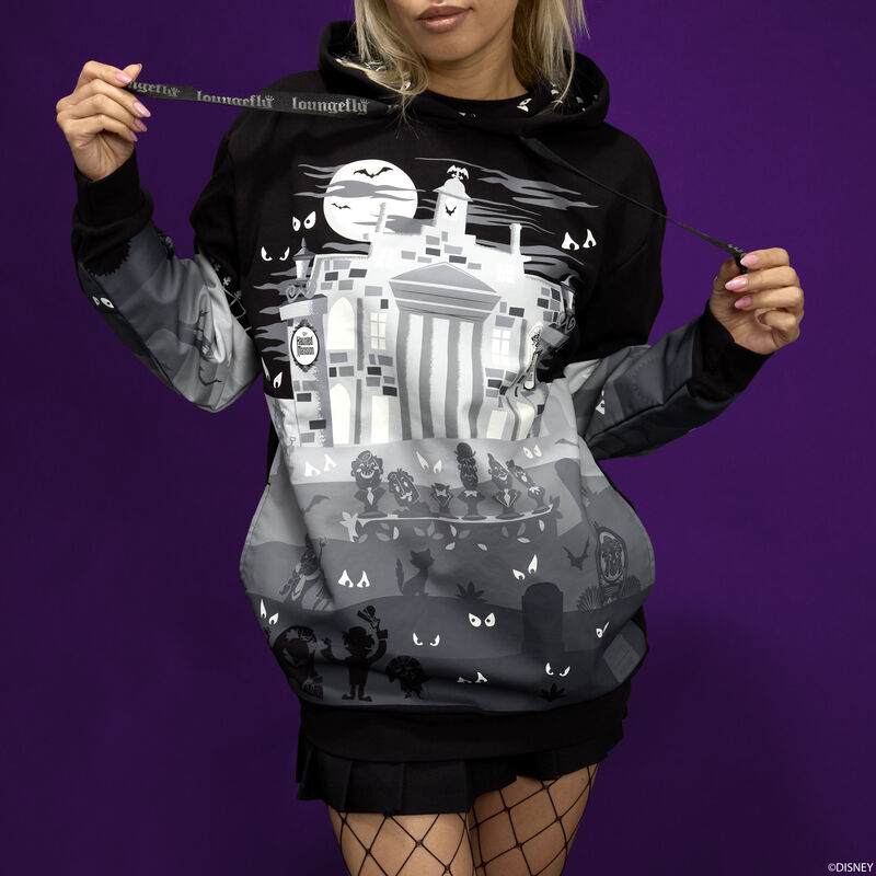 Woman standing against a purple background wearing the Loungefly Haunted Mansion hoodie, which is a black hoodie and has gray and white imagery of the Haunted Mansion on it. 