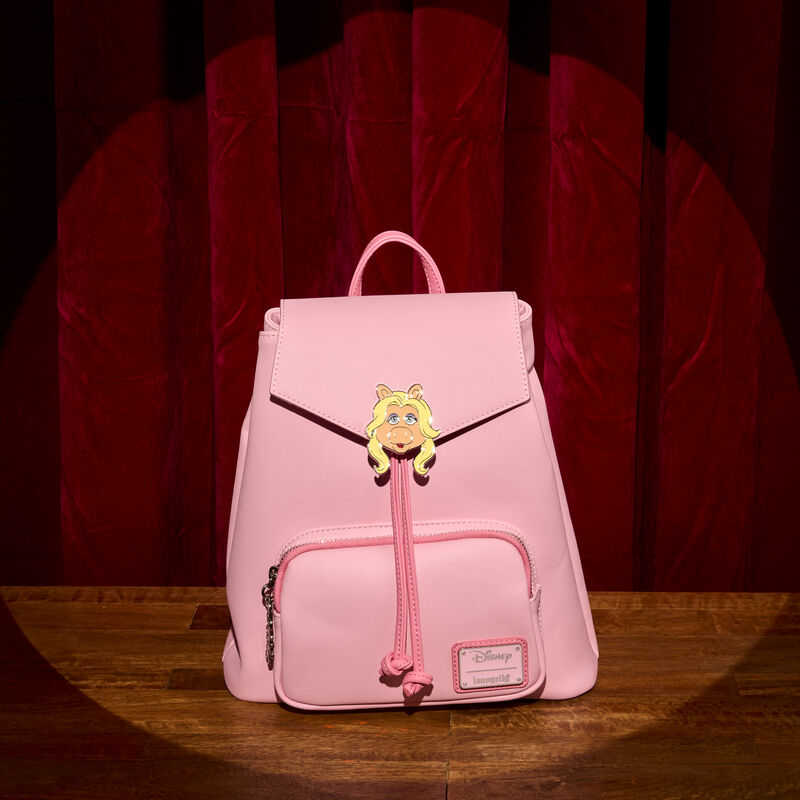 Pink Loungefly Miss Piggy mini backpack with drawstrings and a small front pocket. There’s a Miss Piggy enamel charm on the front flap. It sits on a wooden floor with a red theatre curtain behind it. 