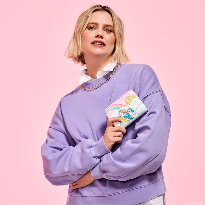 Woman wearing a purple sweatshirt with arms crossed, holding the Loungefly My Little Pony Equestria wallet. She stands against a pink background.  