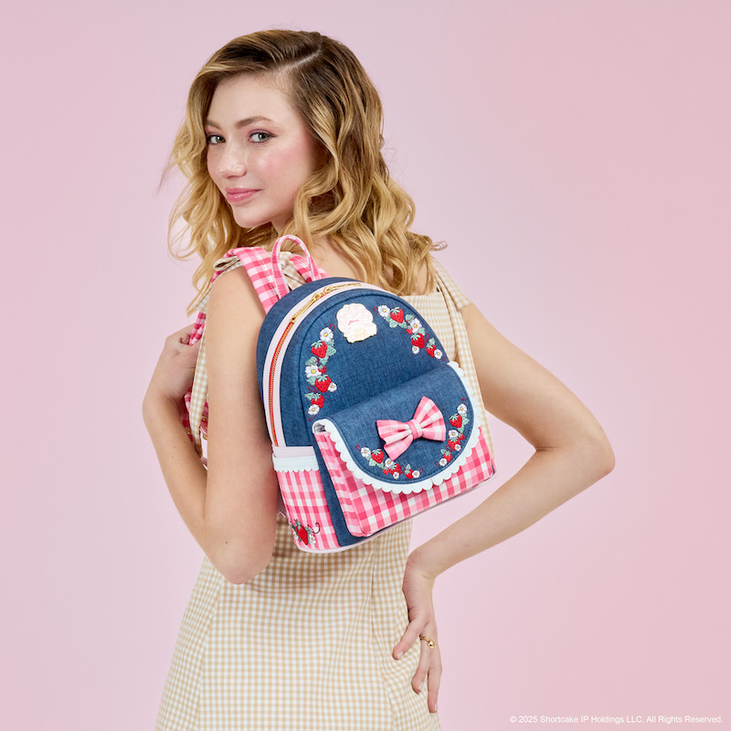 Woman facing away from camera smiling over a shoulder. She wears a yellow-and-white checkered dress and the Loungefly Strawberry Shortcake Springtime Denim mini backpack over a shoulder. She stands in front of a pink background. 