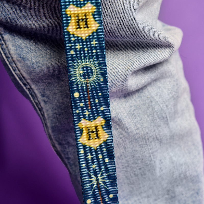 Blue Loungefly leash with a repeating pattern of the Hogwarts crest and wands on it. It's lays against someone's jeans as they hold it, standing against a purple background.