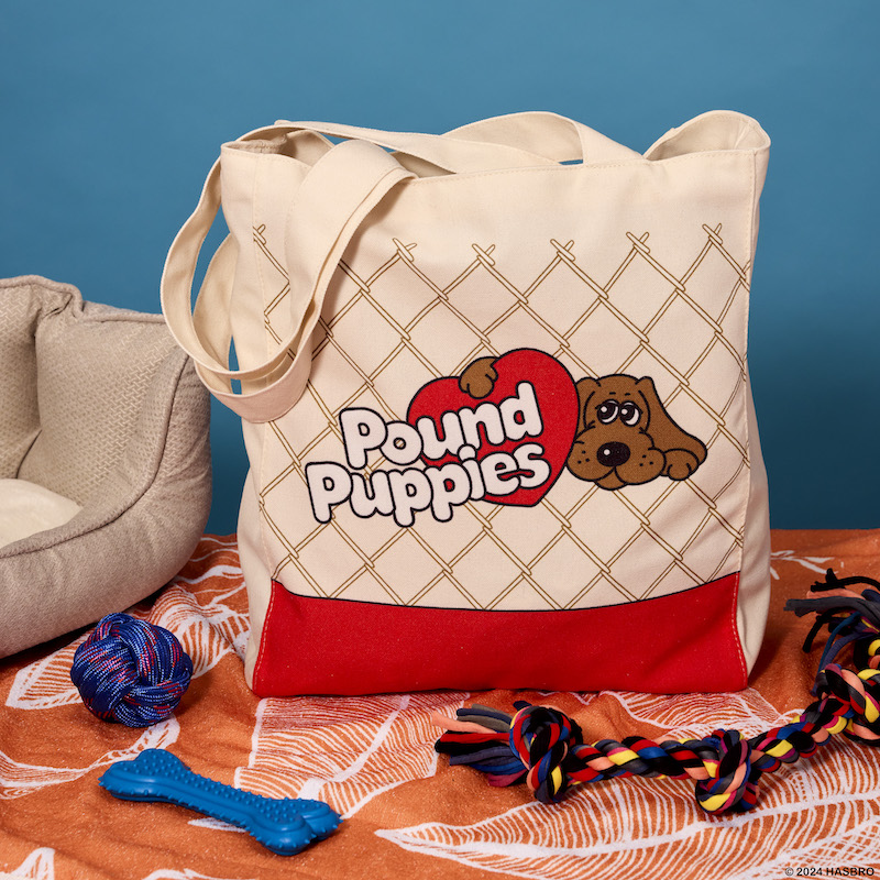 Loungefly Pound Puppies Tote Bag sitting on a table next to a dog bed and dog toys, featuring artwork inspired by the Pound Puppies logo on the front, featuring a brown puppy next to the words "Pound Puppies" over a red heart. 