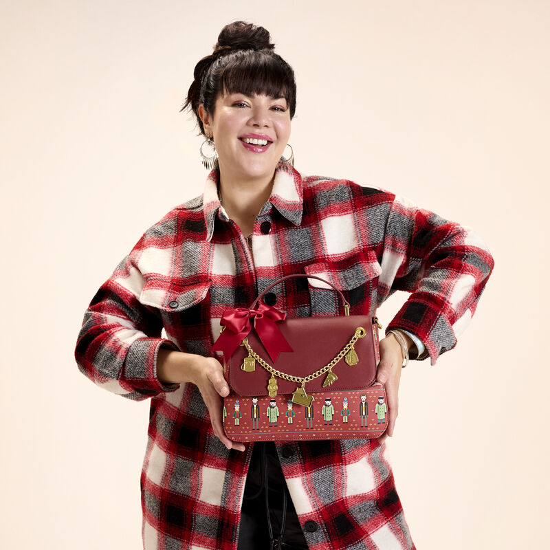 Woman wearing black, white, and red flannel top, holding the Loungefly Home Alone crossbody bag. She stands against a light peach background.
