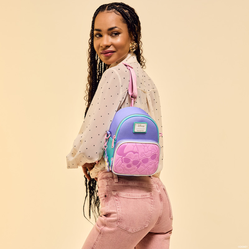 Woman facing away from camera looking over her shoulder and wearing the Loungefly Daisy Duck 85th Anniversary Daisy Duck and Minnie Mouse Convertible Mini Backpack and Crossbody Bag. The woman wears a sheer neutral-toned, black-polka-dot blouse with pink jeans. She stands against a beige background. 