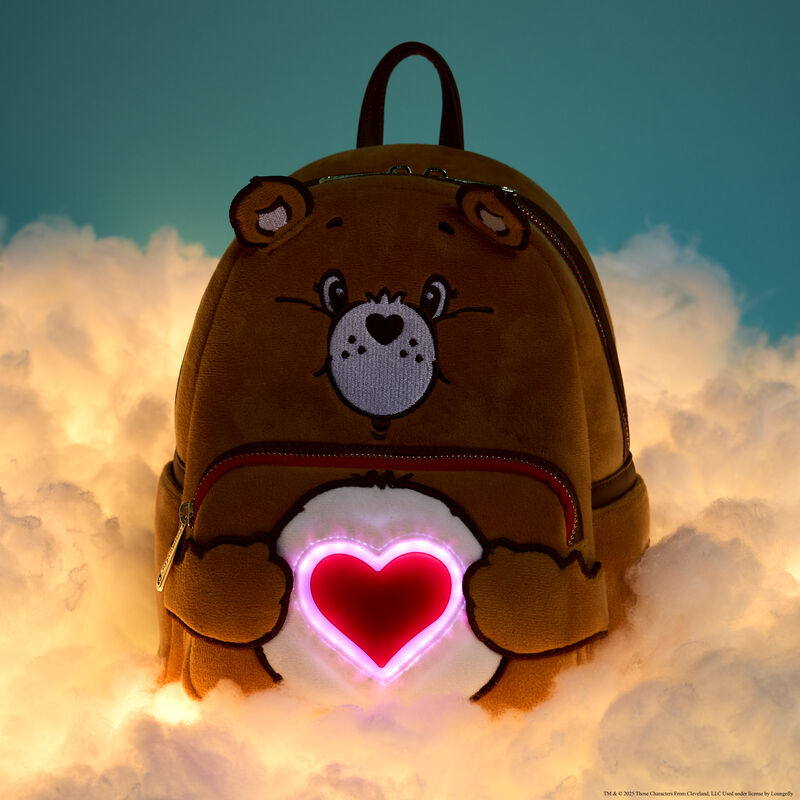 Plush Loungefly Care Bears Tenderheart Bear Mini Backpack with the lights off, showing how the outline of the heart on Tenderheart Bear's stomach lights up! The bag sits on fluffy white clouds.