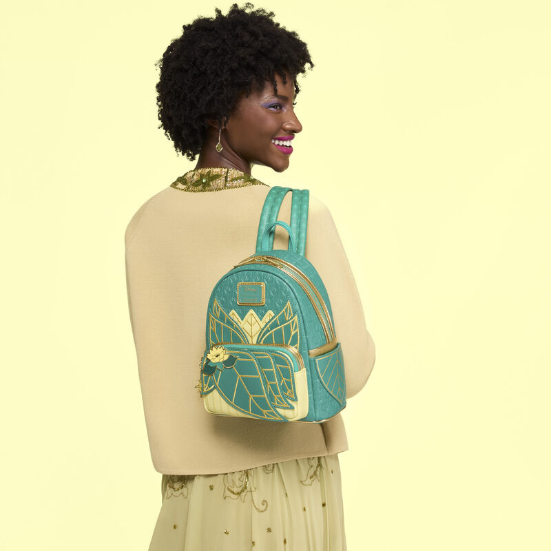 Woman facing away from camera, looking over a shoulder and smiling. She wears the Loungefly Disney Princess Tiana Mini Backpack over a shoulder and stands against a yellow background. 