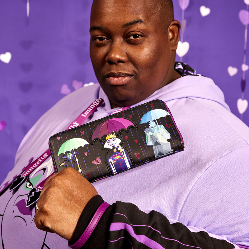 Man holding out the Loungefly Disney Villains Curse Your Hearts Wallet and raising an eyebrow to camera while wearing the Curse Your Hearts hoodie against a purple background with heart-shaped streamers 