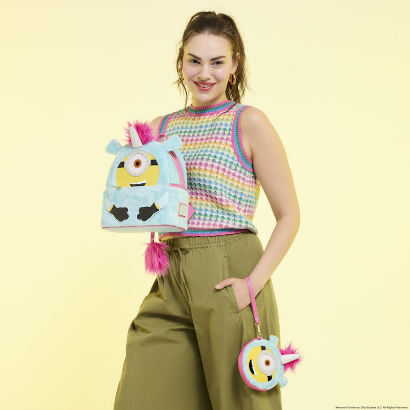 Woman smiling at camera holding the Loungefly Minions Fluffy Unicorn Plush mini backpack and wristlet wallet. She wears a rainbow knit top and green pants. She stands in front of a yellow background. 