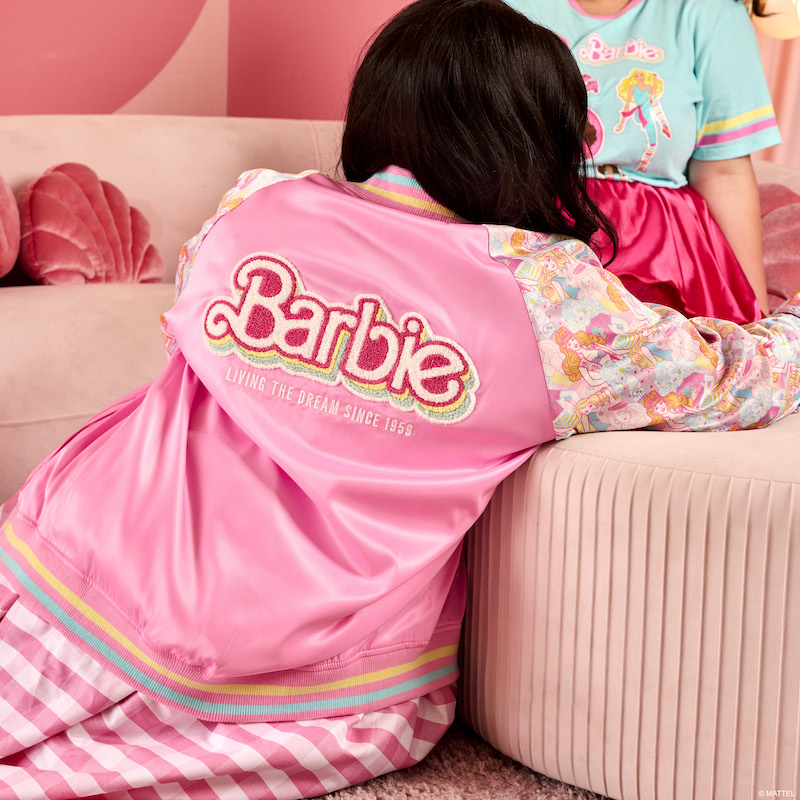 Woman facing away from camera, sitting on the floor wearing the Loungefly Barbie 65th Anniversary Unisex Bomber Jacket. The back of the jacket features a chenille patch that says "Barbie" and underneath, you'll find the words "Living the Dream Since 1959."