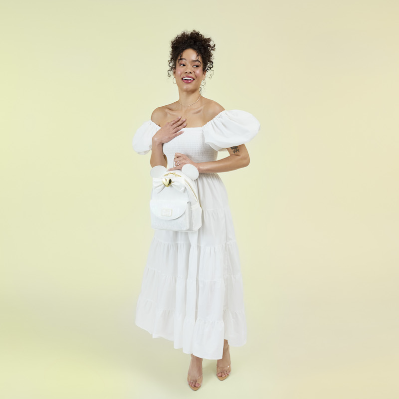 Woman wearing a white off-the-shoulder dress and nude heels, holding the Loungefly Minnie Mouse wedding mini backpack. She stands against a light yellow background. 