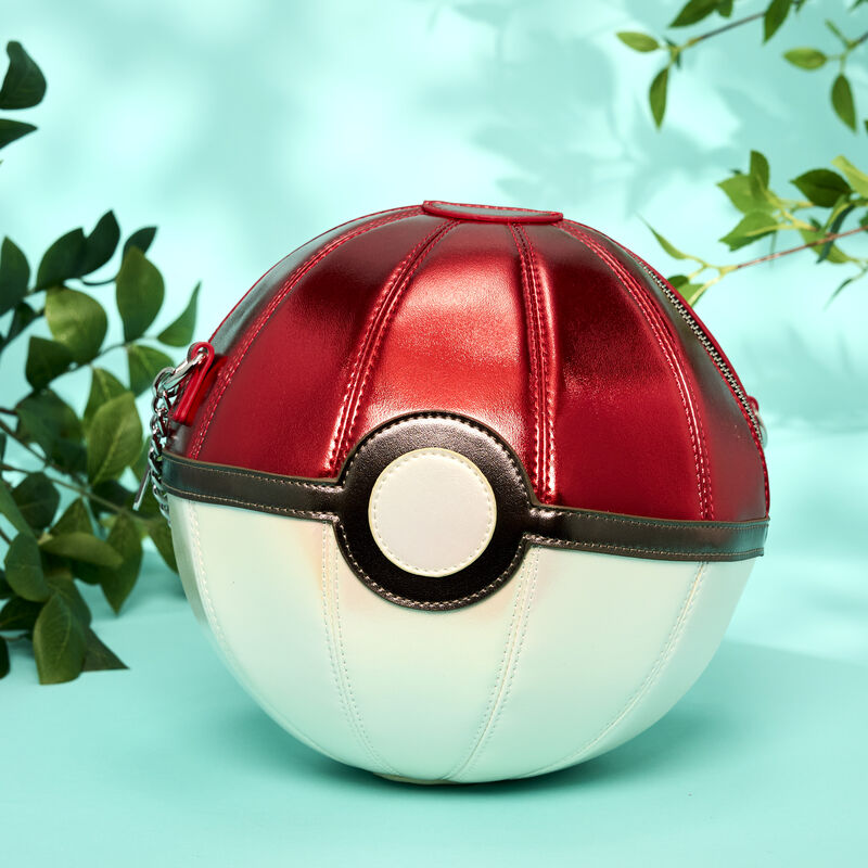 Figural Loungefly crossbody bag in the shape of a red-and-white Pokéball. It sits against a light blue background with green plants around it. 