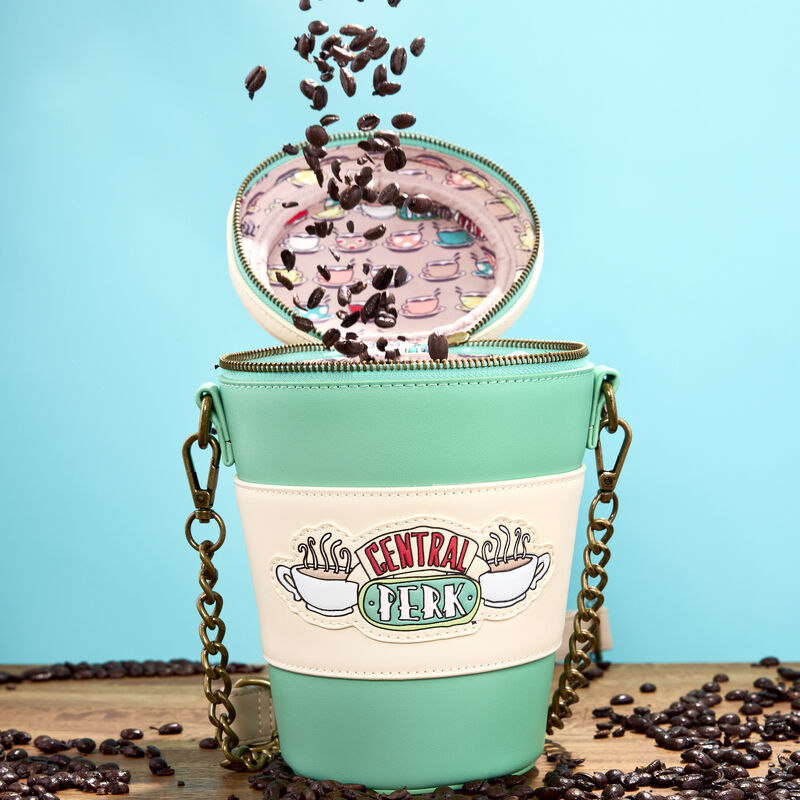 The Central Perk To-Go Coffee Cup Crossbody bag sits on a wooden table against a light blue background, surrounded by coffee beans. Coffee beans fall into the open bag.