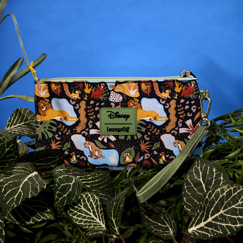 Loungefly Lion King 30th Anniversary All-Over Print wristlet, featuring characters like Simba, Nala, Timon, and Pumbaa amongst various leaves. The wristlet sits on top of green vegetation against a dark blue background. 