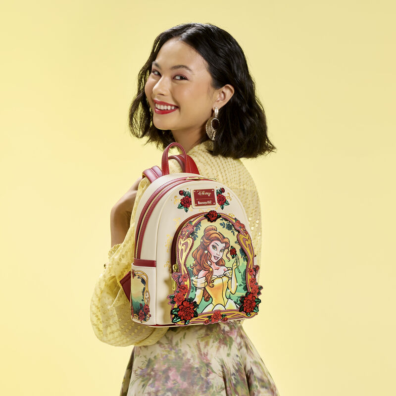 Woman wearing a yellow sweater and floral skirt, looking over a shoulder at camera. She wears the Loungefly Beauty and the Beast Belle Floral Mini Backpack over a shoulder. 