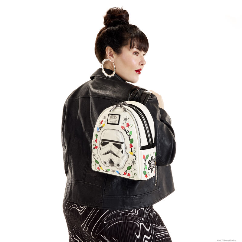 Woman facing away from camera, looking over a shoulder, wearing a black leather jacket and white and black skirt. Over a shoulder, she carries the Loungefly Star Wars Stormtrooper light up mini backpack. She stands against a white background.