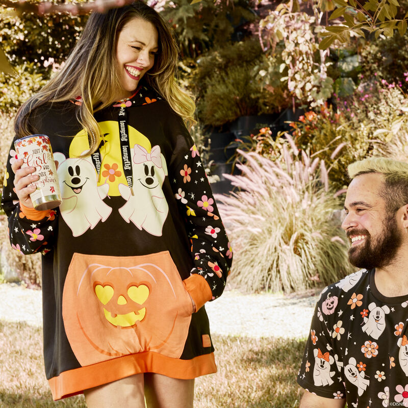 Woman standing outside smiling at a male friend, wearing the Loungefly Mickey and Minnie Floral Ghost Glow Unisex Hoodie, featuring Mickey and Minnie as ghosts floating above a jack-o'-lantern front pocket. 