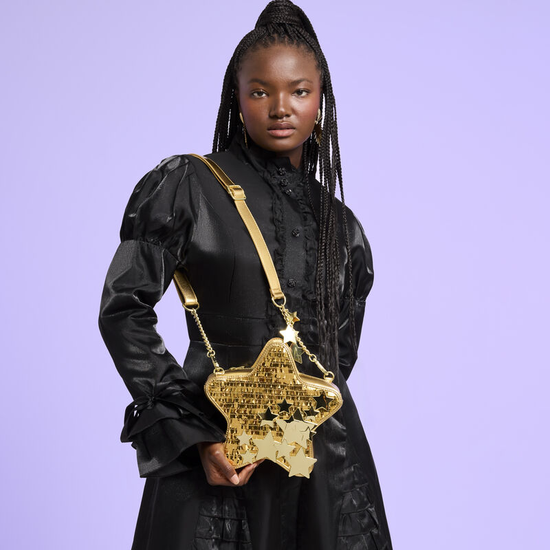 Woman wearing a black dress with the Loungefly Gold Star Sequin Figural Crossbody Bag over a shoulder. She stands against a purple background. 