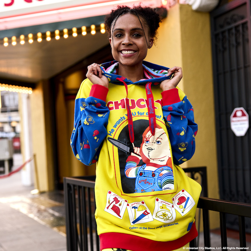 Woman standing outside by an old school movie theatre, wearing the Loungefly Chucky hoodie, featuring the same image as the mini backpack of Chucky with a knife. 