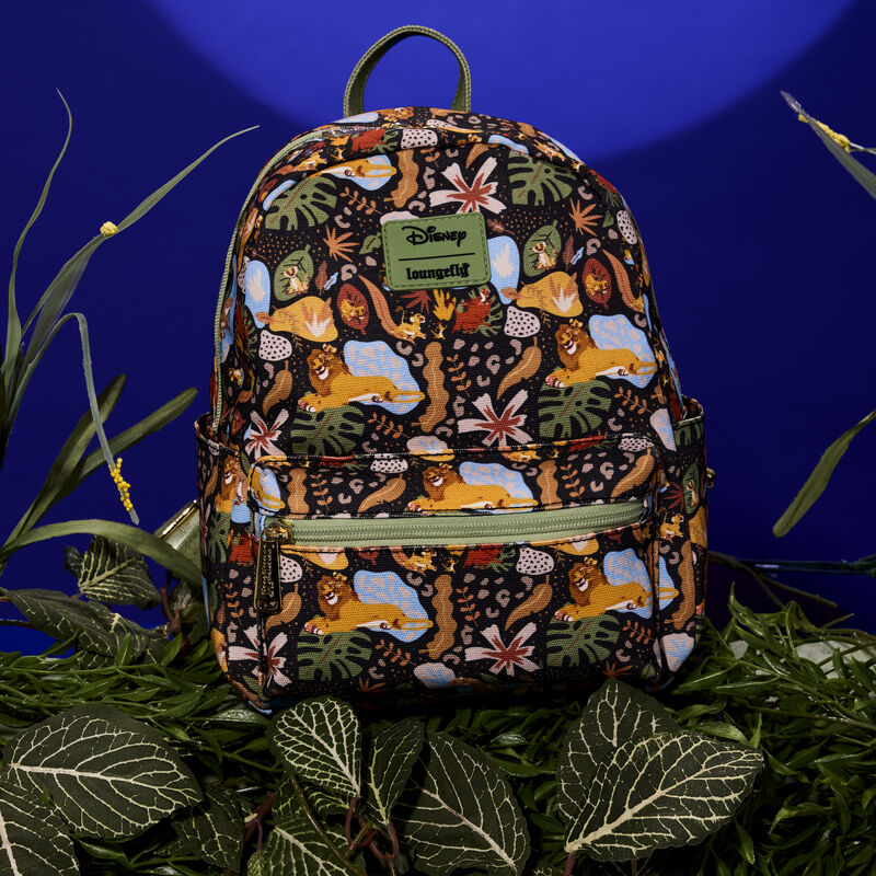 Loungefly Lion King 30th Anniversary All-Over Print mini backpack, featuring an all-over print of Simba, Nala, Timon, and Pumbaa amongst various leaves. The bag sits amongst green vegetation against a dark blue background.