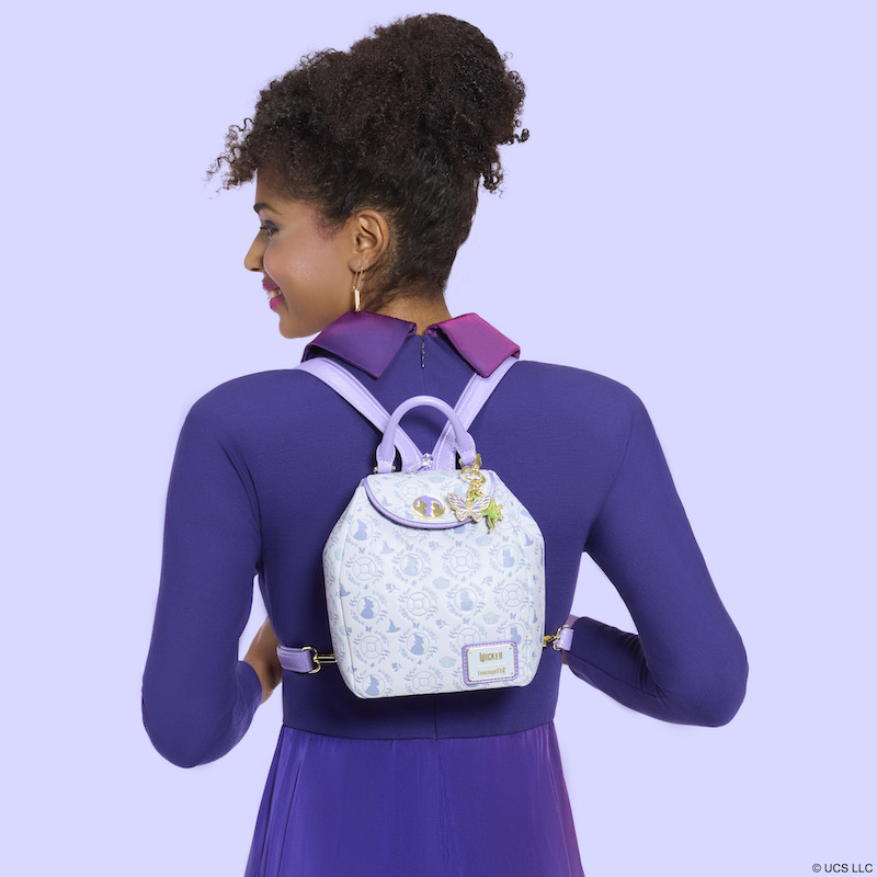 Woman facing away from camera, wearing a purple dress. She smiles over a shoulder and wears the Loungefly Wicked Micro Mini Backpack/Sling as a backpack on her back. She stands against a purple background. 