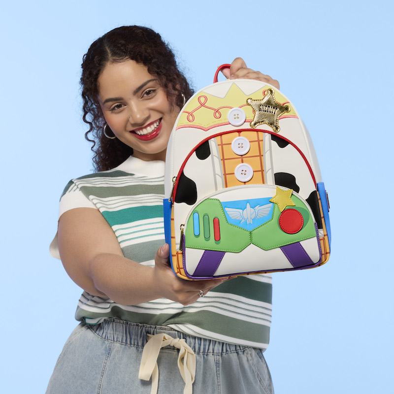 Woman smiling at camera wearing a striped green-and-white top. She holds out the Loungefly Toy Story 5 Triple Pocket backpack out to camera. 
