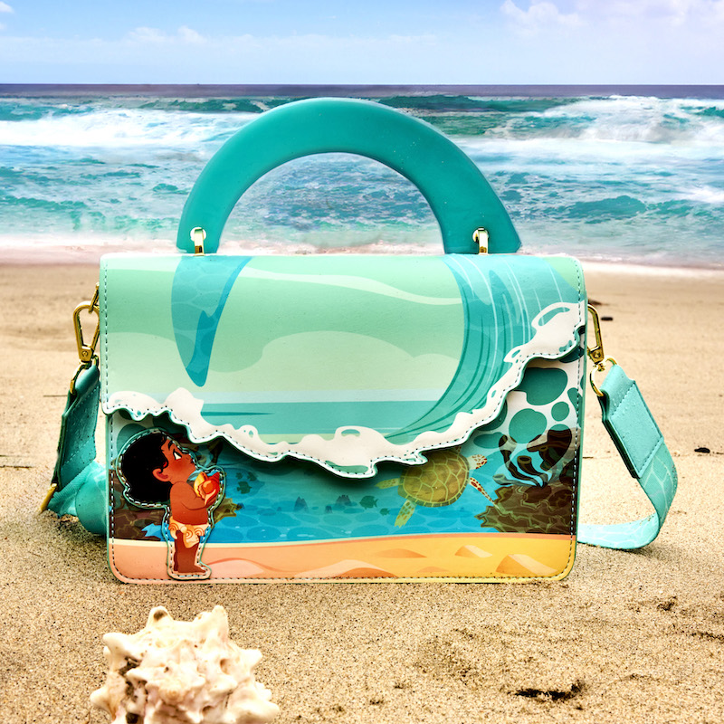 Loungefly Young Moana Wave Crossbody Bag in the sand on the beach with a shell nearby. 