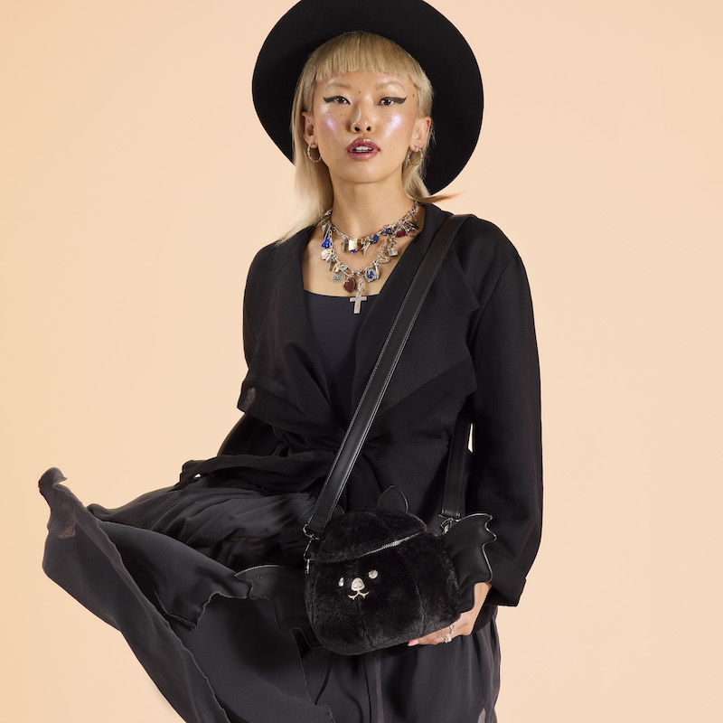Woman wearing a black dress, jacket, and hat, with large statement necklaces, wearing the Loungefly Plush Bat Figural Crossbody Bag over a shoulder. She stands in front of a peach-colored background. 