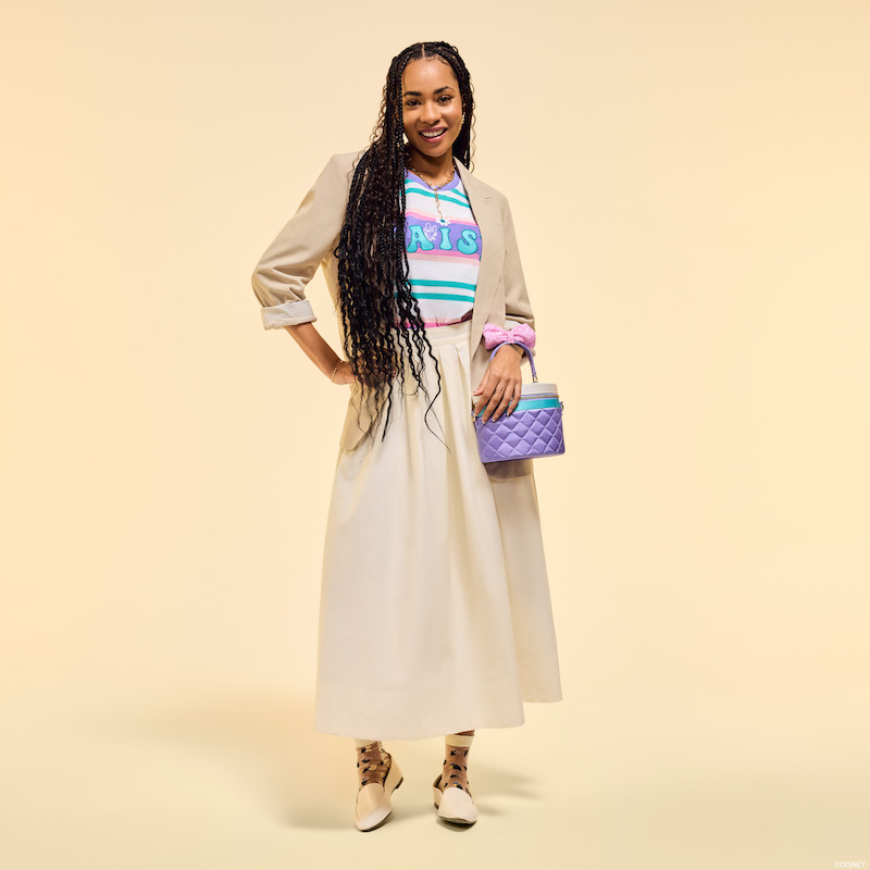 Woman wearing a skirt, Daisy tee, and blazer. She smiles at camera and carries the Loungefly Daisy Duck 85th Anniversary Sequin Quilted Crossbody Bag over one arm. She stands in front of a beige background.