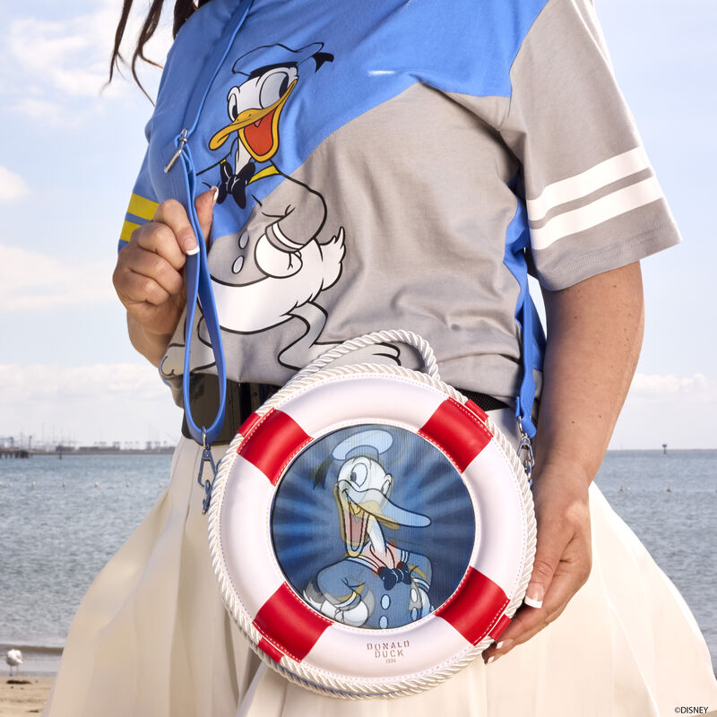 Person at the beach wearing the Loungefly Donald Duck 90th Anniversary Unisex Tee and the lenticular crossbody bag featuring a lenticular panel of Donald Duck within a 3D life preserver ring