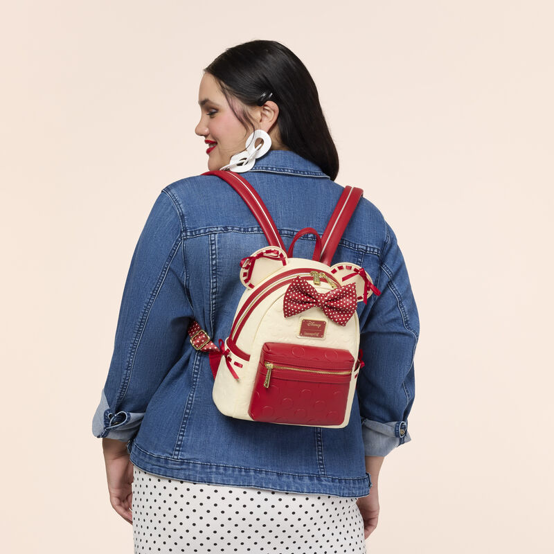 Woman facing away from camera wearing a denim jacket and white skirt with black polka dots. She wears the Loungefly Minnie Mouse Rock the Dots mini backpack. 