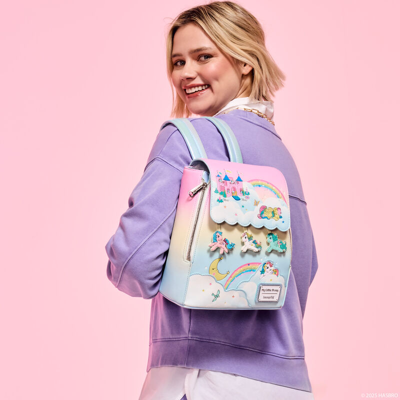 Woman wearing a purple sweater facing away from camera and smiling over a shoulder. She wears the rainbow ombre Loungefly My Little Pony Equestria mini backpack. 