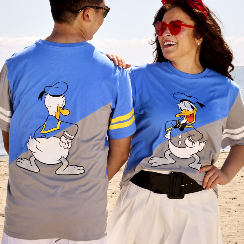 Man and woman standing together on the beach wearing the Loungefly Donald Duck 90th Anniversary Unisex Tee, one facing camera and one facing away from camera to show off the front and back of the shirt, which features Donald Duck in the center of it 