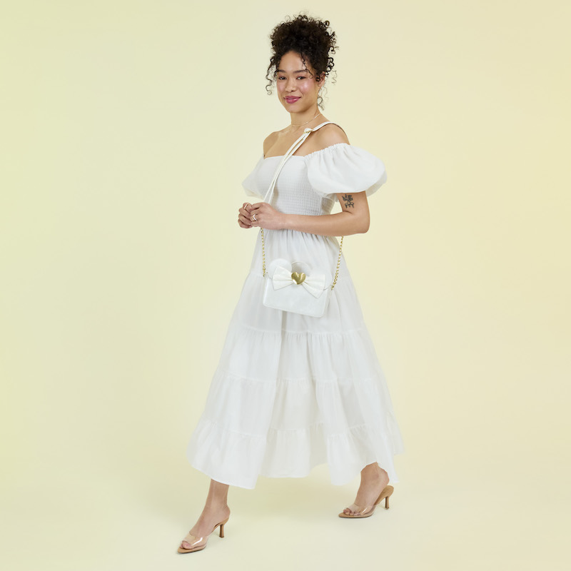 Woman wearing a white off-the-shoulder dress and nude heels carrying the Loungefly Disney Minnie Mouse Iridescent Lace Wedding Crossbody. She stands against a light yellow background. 